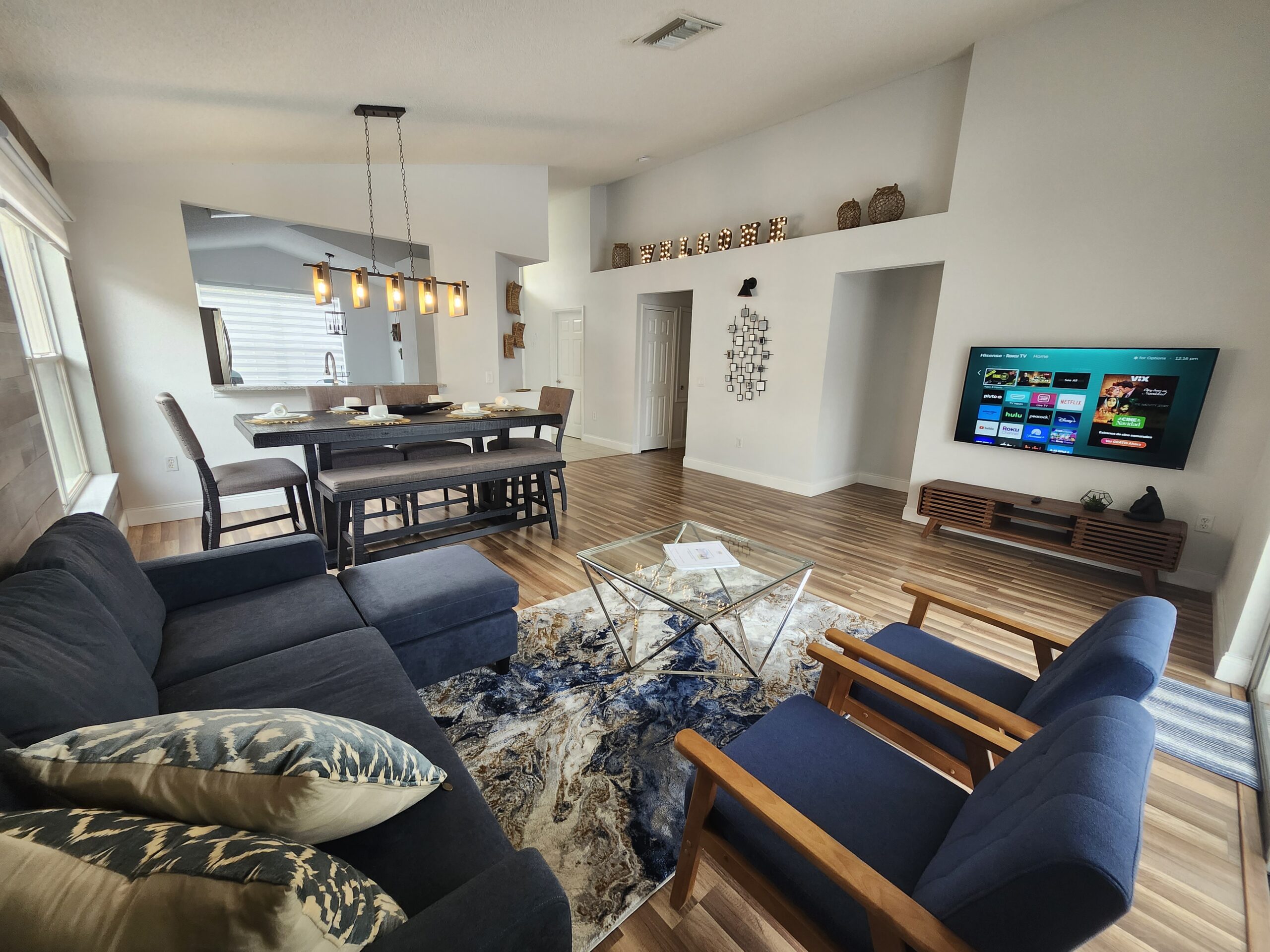 Finished open-concept vacation rental living and dining room in Orlando with a lit WELCOME marquee sign, blue sectional and wall-mounted TV